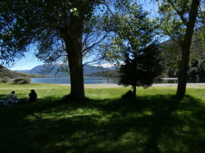 Just arrived at Loch Katrine to a stunning day. Under some trees with some people sitting under the shade to the left. Loch Katrine out past that with mountains on the side